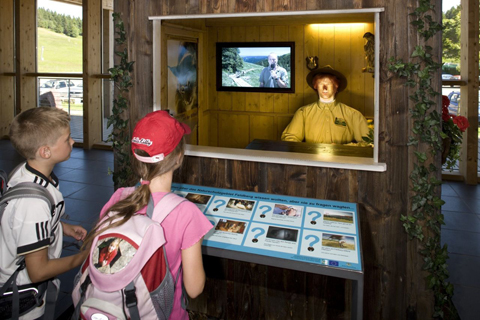 Kleine Holzhütte mit Glasscheibe, hinter der eine Ranger-Puppe sitzt, an der Innenwand ist ein Monitor angebracht. Fragen kann man über Tasten vor der Scheibe stellen.
