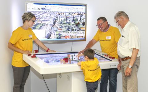 Auf dem Foto stehen drei erwachsene Personen mit einem Jungen vor einer 3D-Visualisierung einer Stadt, die auf einem Bildschirm gezeigt wird.