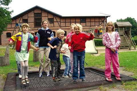 8 größere und kleinere Kinder hüpfen auf einem Trampolin. Im Hintergrund ist ein Gebäude aus roten Backsteinen zu sehen.