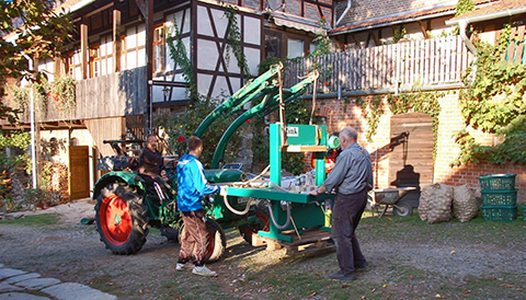 <p>
	2 Männer und 1 Junge bringen die Saftpresse mit einem Traktor an ihren neuen Platz. Im Hintergrund ein Teil der Hofgebäude aus Ziegeln und Fachwerk</p>

